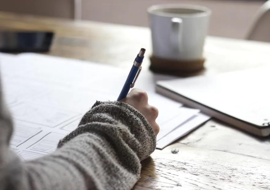 A hand writing notes beside a coffee cup at a desk